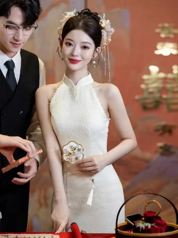 Elegant bride wearing a white halter cheongsam with delicate pearl decorations and a traditional floral headpiece, standing gracefully next to a groom in a black vest during a Chinese wedding.